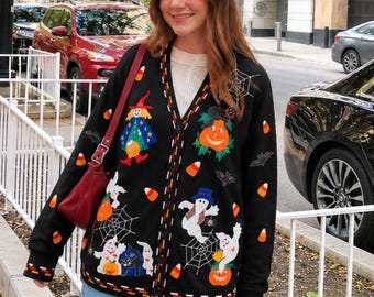 May include: A black V-neck cardigan featuring colorful Halloween-themed embroidery including ghosts, a witch, a pumpkin, spiderwebs, bats, and candy corn. The cardigan has a button-down front and a patterned trim along the cuffs and hem, worn over a white top.