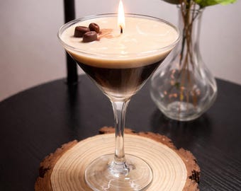 May include: A martini glass candle with a layered appearance, featuring a dark brown base and a creamy white top. The candle is lit, with a small flame, and is decorated with three dark brown coffee bean-shaped wax accents on the surface.