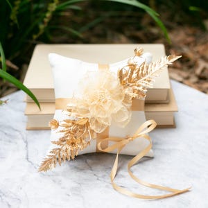 May include: A square, white ring pillow with a gold ribbon and a gold floral arrangement. The pillow is decorated with a large, layered flower and gold-painted leaves. A gold ribbon is tied in a bow.
