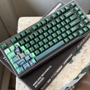 May include: A wireless mechanical keyboard with green keycaps and a dark gray frame. The keycaps have white lettering. The space bar features a comic book character. The keyboard is on top of a black box with the words "WIRELESS MECHANICAL KEYBOARD".