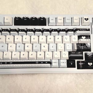 May include: A white mechanical keyboard with custom keycaps. The keycaps feature a black and white color scheme with decorative patterns, including bows, hearts, and lace designs. The keyboard has a standard layout.