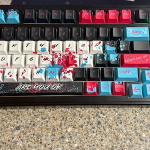 May include: A mechanical keyboard with a black frame and custom keycaps featuring a paint splatter design in red, blue, and white. The spacebar displays the text ARE YOU OK in white lettering, and various keys feature red, blue, and black accents.