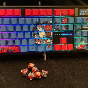 May include: A black and red mechanical keyboard with blue keys featuring Spider-Man graphics. The keyboard has red keycaps on the top row and a small display screen. Several red and gray key switches are in front of the keyboard.