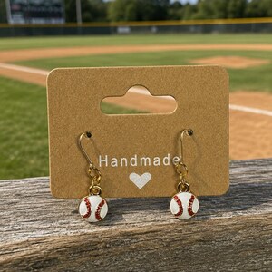 May include: A pair of baseball-themed earrings. Each earring features a small, white baseball with red stitching and gold-colored hooks. The earrings are displayed on a brown card with the word "Handmade" and a heart symbol.