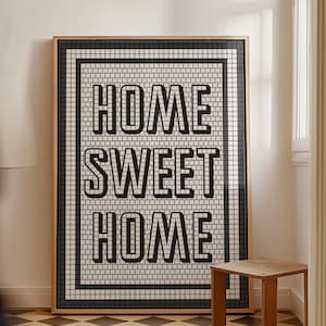 May include: A framed wall art piece with the words "HOME SWEET HOME" in bold, black letters. The artwork has a black and white tile design and a gold frame. A small wooden stool sits nearby on a checkered floor.