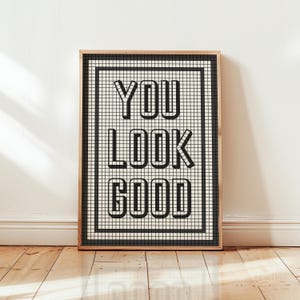 May include: A framed print with the words "YOU LOOK GOOD" in bold, black letters. The text is centered on a cream-colored background with a black grid pattern. The frame is a light brown color, and the print is leaning against a white wall.