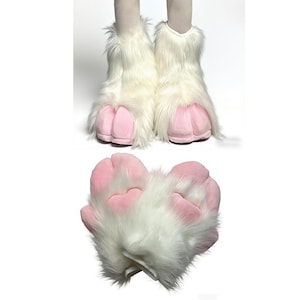 Op de afbeelding: Witte imitatiebont laarzen met roze pootkussentjes. De laarzen hebben een donzige textuur en zijn ontworpen om op dierenpoten te lijken. De roze kussentjes hebben een zachte, afgeronde vorm. Er wordt ook een apart pootkussentje getoond.