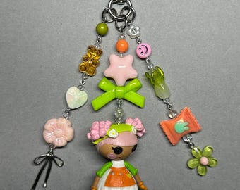 May include: A keyring featuring a silver heart-shaped clasp with three dangling beaded strands. The centre strand includes a pink star, a lime green bow, and a small doll figure with pink hair, an orange dress, and white apron.