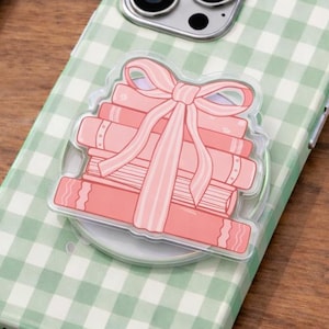 Op de afbeelding: Groen en wit geruit telefoonhoesje met een stapel roze boeken en een roze strik. De telefoonhoes heeft een doorzichtige pop-up grip aan de achterkant.