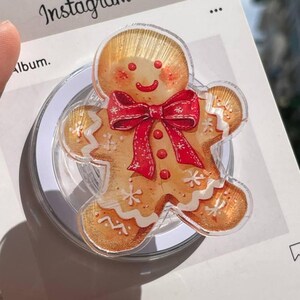 Könnte beinhalten: Ein Handyhalter in Form eines Lebkuchenmannes mit roter Schleife und weißen Akzenten. Der Halter ist an einer transparenten, kreisförmigen Basis befestigt. Das Wort "Instagram" ist im Hintergrund zu sehen.