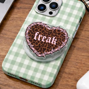 May include: A green and white gingham patterned phone case with a leopard print heart-shaped phone grip that says "freak" in pink lettering. The phone has three camera lenses.