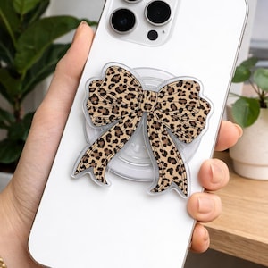 May include: A white smartphone with a leopard print bow phone grip attached. The bow is tan with dark brown spots. The phone has a triple-lens camera and is held in a hand with a gold bracelet.