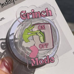 May include: A clear, circular pop socket with a Grinch-themed design. The design features the words "Grinch Mode" in pink, along with a green Grinch hand turning an "ON/OFF" switch. The pop socket is attached to a white backing.