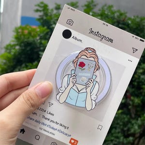 May include: A clear phone grip featuring a cartoon illustration of a woman holding a book with a rose and candelabra. The grip is set against a white Instagram-themed background with text. The woman is wearing a light blue dress.