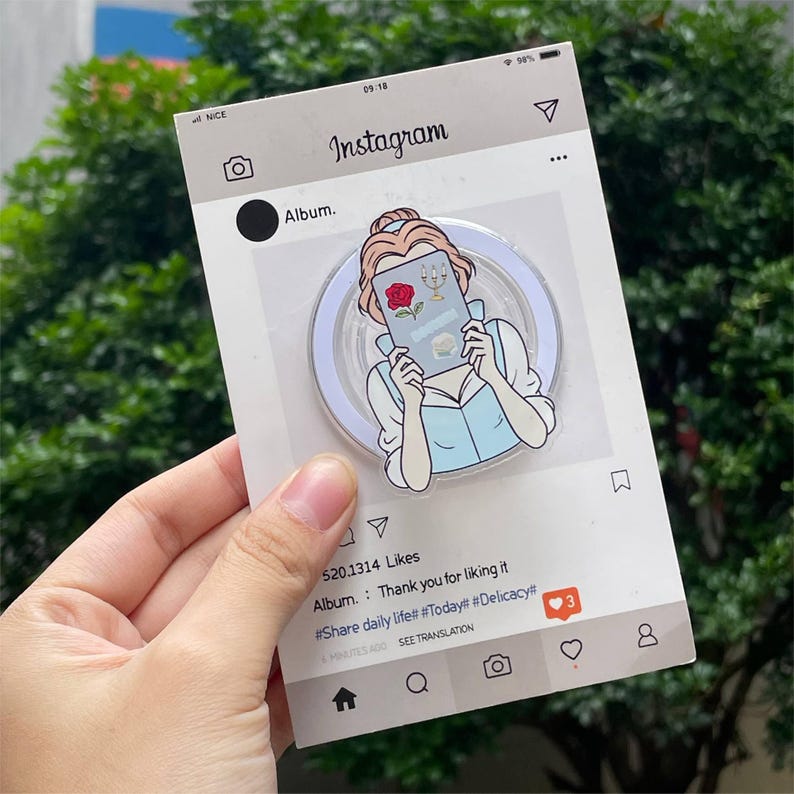 May include: A clear phone grip featuring a cartoon illustration of a woman reading a book, with a rose and candelabra on the cover. The grip is set against a white background with an Instagram-themed design, including the text "Instagram" and "Album."
