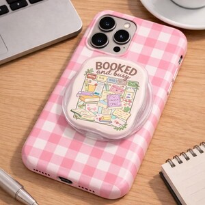May include: A pink and white gingham patterned phone case with a pop-up grip. The grip features the text "booked and busy" with a colorful illustration of books, notes, and a calendar. The phone case is on a wooden surface.