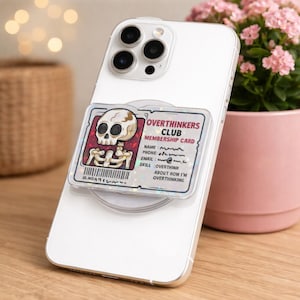 May include: A white smartphone with a clear phone grip featuring an "Overthinkers Club" membership card design. The card has a skull graphic and text that reads "Overthinkers Club Membership Card".