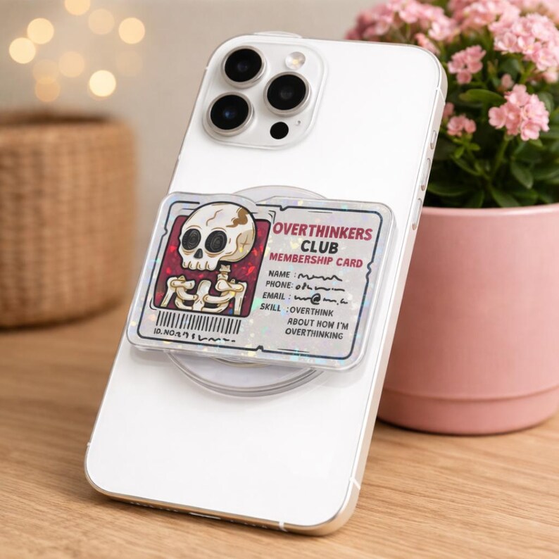 May include: A white smartphone with a clear phone grip featuring an "Overthinkers Club" membership card design. The card has a skull graphic and text that reads "Overthinkers Club Membership Card".