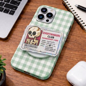 May include: A green and white gingham patterned phone case with a pop-out grip. The grip features an "Overthinkers Club" membership card design with a skull and skeleton graphic. The phone is on a wooden surface.