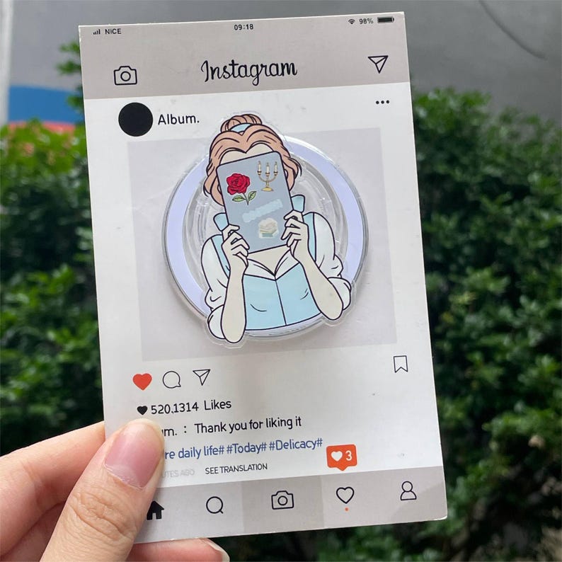 May include: A clear acrylic phone grip featuring a cartoon illustration of a woman holding a book with a rose and candelabra. The design is set against a white background with an Instagram-themed frame, including a like count and comments.