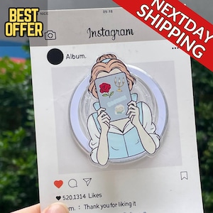 以下が含まれることがあります： 赤いバラとろうそくのデザインが施された本を持つ女性の漫画イラストが描かれた透明な電話グリップ。 グリップは、テキスト「Instagram」を含むInstagramをテーマにしたフレームの白い背景に設定されています。