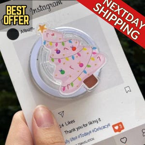 Puede incluir: Un soporte para teléfono con forma de árbol de Navidad rosa con luces de colores, adornos y una estrella. El soporte está sobre un fondo blanco con las palabras "BEST OFFER" y "NEXTDAY SHIPPING".