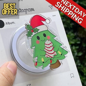 May include: A Christmas tree-shaped phone grip with a Santa hat, holly, and string lights. The green tree has a smiling face and a gingerbread cookie design. The grip is attached to a white and clear circular base.
