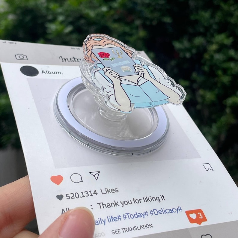 May include: A clear acrylic phone grip featuring a cartoon illustration of a woman reading a book, with a red rose and a golden candelabra. The grip is mounted on a white and silver circular base, presented on a social media-themed card.