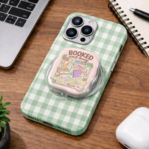 May include: A green and white gingham patterned phone case with a pop-up grip. The grip has the words "booked and busy" and a colorful illustration of books and a calendar. The phone case is on a wooden surface.