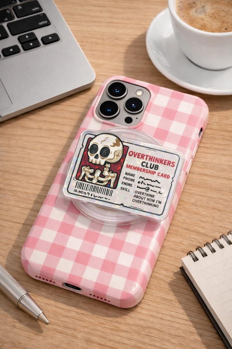 May include: Pink gingham iPhone case with a pop-up grip featuring an "Overthinkers Club" membership card design. The card has a skeleton graphic and text. A laptop, pen, and coffee cup are in the background.