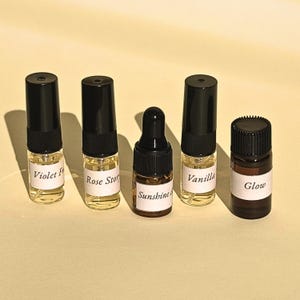 Könnte beinhalten: Fünf kleine Glasflaschen mit schwarzen Verschlüssen und weißen Etiketten. Vier Flaschen sind klar mit Sprühaufsatz und beschriftet mit Violet F, Rose Story, Sunshine und Vanilla. Die fünfte Flasche ist aus Braunglas mit Pipette und trägt die Aufschrift Glow.