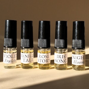May include: Five miniature perfume bottles with black lids and white labels. The labels read "VIOLET Fraîche", "VANILLA TALE", "ROSE STORY", "IRIS SONG", and "NIGHT Blooms". Each bottle contains a pale yellow liquid.