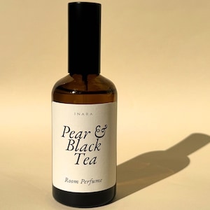 May include: A brown glass spray bottle with a black cap and a cream-colored label. The label features the text INARA, Pear & Black Tea, and Room Perfume in black font. The bottle has a cylindrical shape and a smooth, dark glass finish.