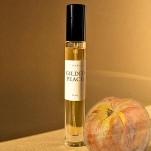 May include: A clear glass perfume roller bottle with a black cap and a white label reading INARA GILDED PEACH 10 ML. The bottle contains a golden liquid and is positioned next to a round peach with yellow, orange, and pink skin tones.