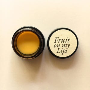 Könnte beinhalten: Geöffnetes Lippenbalsam-Glas mit gelb-oranger Farbe, neben dem Deckel. Der Deckel ist weiß mit dem Schriftzug "Fruit on my Lips" in schwarzer Schrift. Das Glas ist dunkelbraun mit schwarzem Rand.
