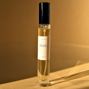 Peut inclure: Un flacon de parfum en verre transparent avec un bouchon noir et une étiquette rectangulaire blanche. L'étiquette porte le texte INARA, DARK et 10 ML en noir. Le flacon contient un liquide ambré clair et un tube pulvérisateur interne.