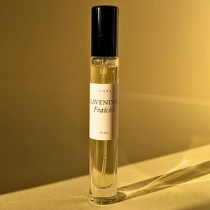 May include: A clear glass perfume spray bottle with a black cap and a white label. The label features the text INARA, LAVENDER Fraîche, and 10 ml. The bottle contains a pale liquid and is positioned vertically against a warm, golden-toned background.