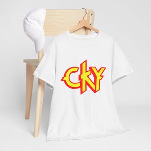 May include: White t-shirt with the yellow and red text "CKY" on the front. The t-shirt is hanging on a wooden hanger. A white baseball cap rests on the back of a wooden chair.