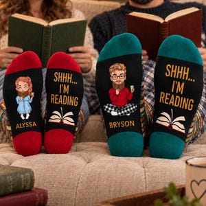 May include: Two pairs of socks featuring black soles with the text SHHH... I'M READING and a graphic of an open book. One pair has red cuffs and a cartoon illustration of a person named ALYSSA, while the other has teal cuffs and a cartoon of BRYSON.