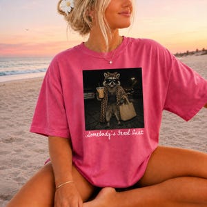 May include: A pink short-sleeve t-shirt featuring a graphic of a raccoon wearing sunglasses, a leopard print coat, and a headscarf while holding a coffee cup and a handbag. The text below the graphic reads, Somebody's Feral Aunt.