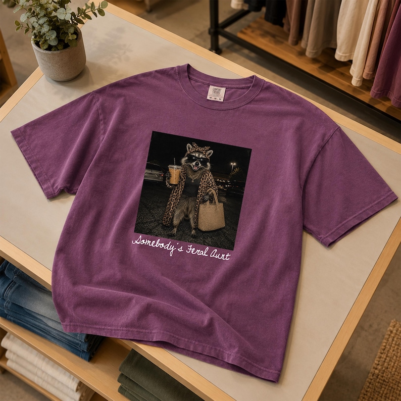 May include: A purple short-sleeve t-shirt featuring a graphic of a raccoon wearing sunglasses, a leopard print coat, and a headscarf while holding a coffee cup and a tote bag. The text below the graphic reads, "Somebody's Feral Aunt" in white script.