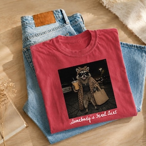 May include: A red t-shirt featuring a graphic of a raccoon wearing sunglasses, a leopard print robe, and a gold chain, while holding a beverage and a tote bag. The text "Somebody's Feral Aunt" is printed in white script below the graphic.