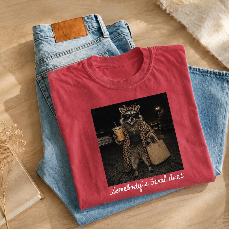 May include: A red t-shirt featuring a graphic of a raccoon wearing sunglasses, a leopard print robe, and a gold chain, while holding a beverage and a tote bag. The text "Somebody's Feral Aunt" is printed in white script below the graphic.