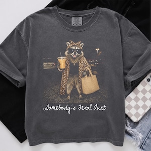 May include: A charcoal gray short-sleeve t-shirt featuring a graphic of a raccoon wearing sunglasses, a leopard print headscarf, and a matching coat. The raccoon holds an iced coffee and a tan tote bag. The text reads, "Somebody's Feral Aunt."
