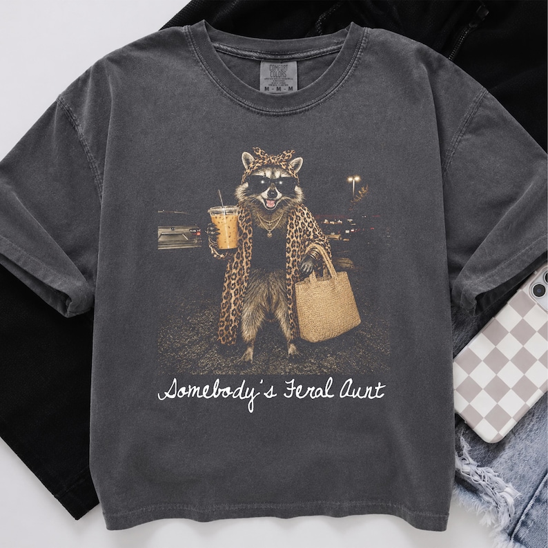 May include: A charcoal gray short-sleeve t-shirt featuring a graphic of a raccoon wearing sunglasses, a leopard print headscarf, and a matching coat. The raccoon holds an iced coffee and a tan tote bag. The text reads, "Somebody's Feral Aunt."