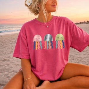 Puede incluir: Una camiseta de manga corta de color rosa brillante con un estampado gráfico de tres medusas. Las medusas tienen capuchones con patrones de cuadros vichy rosas, rayas azules y cuadros vichy verdes, con tentáculos ondulados y ojos en forma de X.