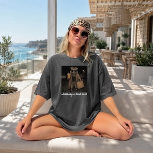 May include: A charcoal gray oversized t-shirt featuring a graphic print of a raccoon wearing a leopard print coat and sunglasses while holding a coffee cup and a tote bag. The text below the raccoon reads, Somebody's Feral Aunt.