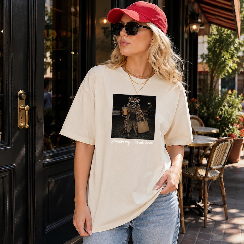 May include: A cream-colored t-shirt featuring a graphic print of a raccoon wearing sunglasses, a leopard print coat, and holding a coffee cup and a handbag. Below the graphic, the text reads Somebody's Feral Aunt in a white cursive script font.