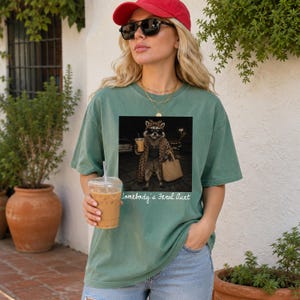 May include: A sage green short-sleeve t-shirt featuring a graphic print of a raccoon wearing a leopard print coat, sunglasses, and a headscarf, while holding a coffee cup and a handbag. The text below the graphic reads, Somebody's Feral Aunt.