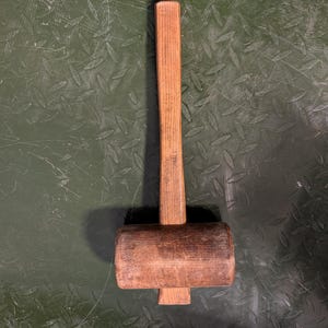 May include: A wooden mallet featuring a cylindrical head and a straight handle. The tool displays a natural brown wood grain texture with visible signs of wear and use, including surface scuffs and indentations on the head and handle.